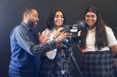 A middle aged teacher standing behind a video camera with two secondary aged students in school uniforms smiling.